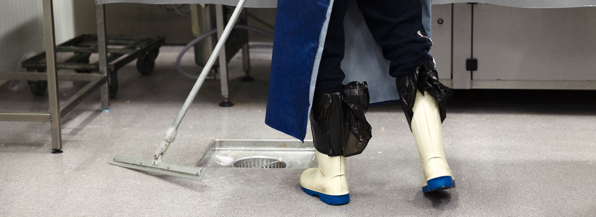 Teollisuussiivoojan jalat kumisaappaissa ja suojavarusteissa pesemässä tuotantotilan lattiaa lastalla lattiakaivon vieressä