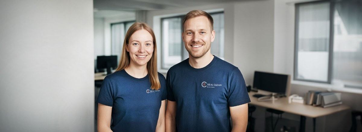 Real Clean -siivouspalvelun kaksi hymyilevää ammattilaista tummansinisessä työasussa modernissa toimistossa